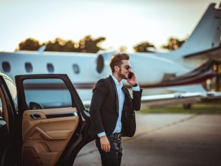 Young rich businessman talking on a mobile phone while getting out of a luxurious car parked next to a private airplane on a tarmac. - Sharon Carr Travel